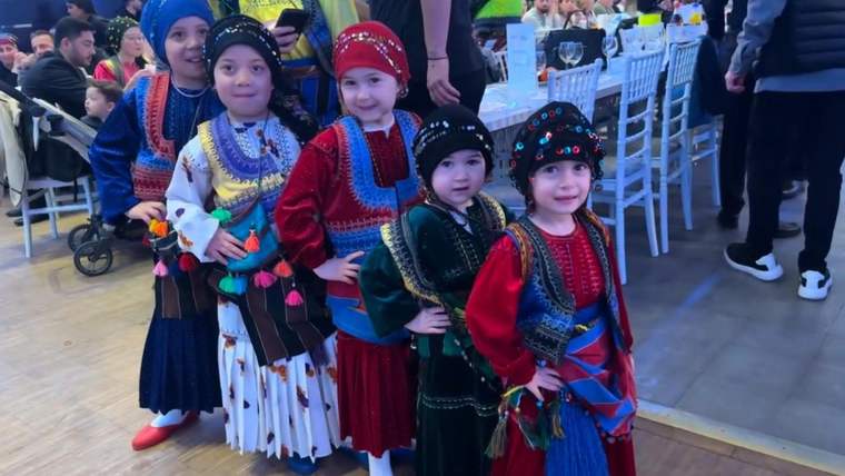 Geleneksel kıyafetler ve başlıklar giyen beş çocuk, kapalı bir mekânda yan yana duruyor.