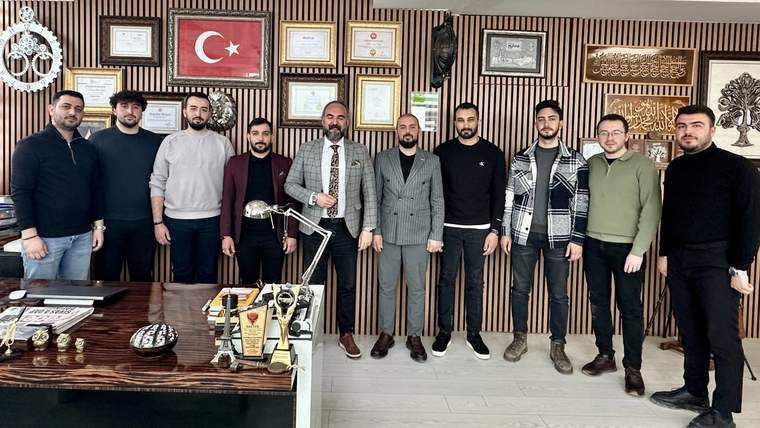 Bir grup erkek, ofis ortamında bir arada duruyor; arka planda Türk bayrağı ve çeşitli sertifikalar var.