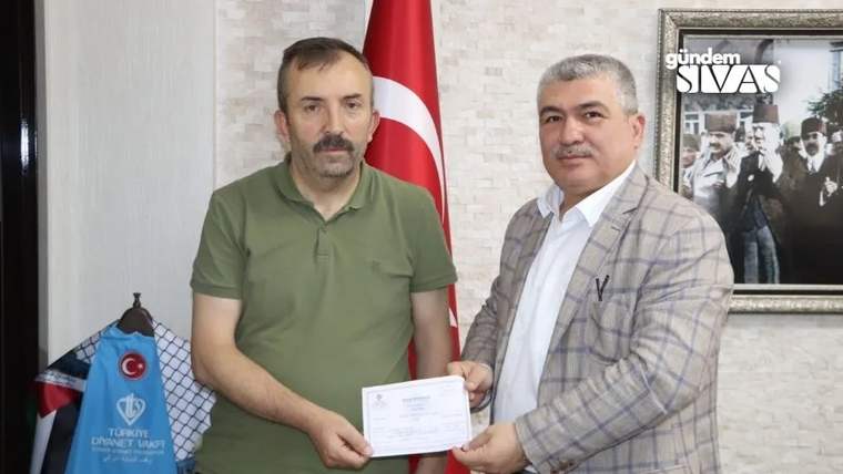 Bir kişi, diğerine bağış belgesi verirken el sıkışıyor; arka planda Türk bayrağı var.