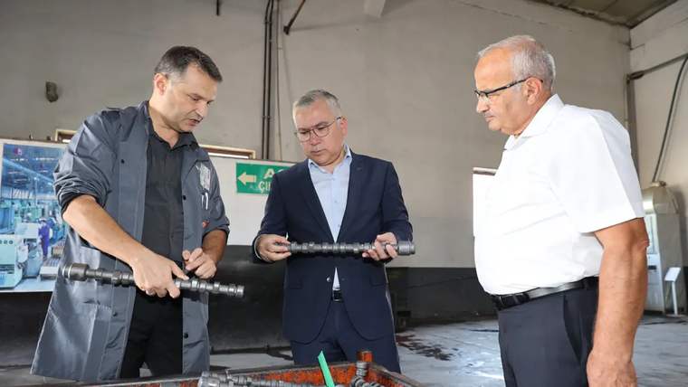 Atölyede üç erkek, ellerindeki metal parçayı incelerken önlerinde sıralanmış metal parçalar bulunuyor.