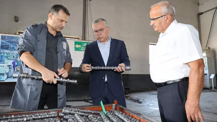 Atölyede üç erkek, ellerindeki metal parçayı incelerken önlerinde sıralanmış metal parçalar bulunuyor.