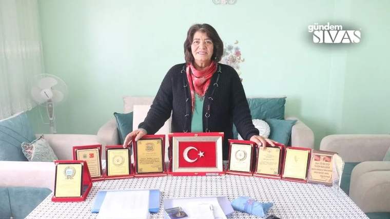 Bir kadın, masadaki üzerinde Türk bayrağı bulunan plaket ve çeşitli belgelerin arkasında duruyor.