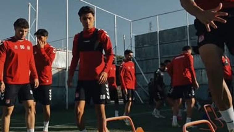 Kırmızı formalar giyen futbolcular, çitle çevrili sahada turuncu engeller arasında antrenman yapıyor.
