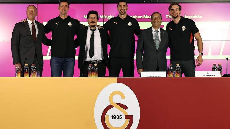 Galatasaray logosu bulunan masanın arkasında yan yana duran altı erkek ve masadaki su şişeleri görülüyor.