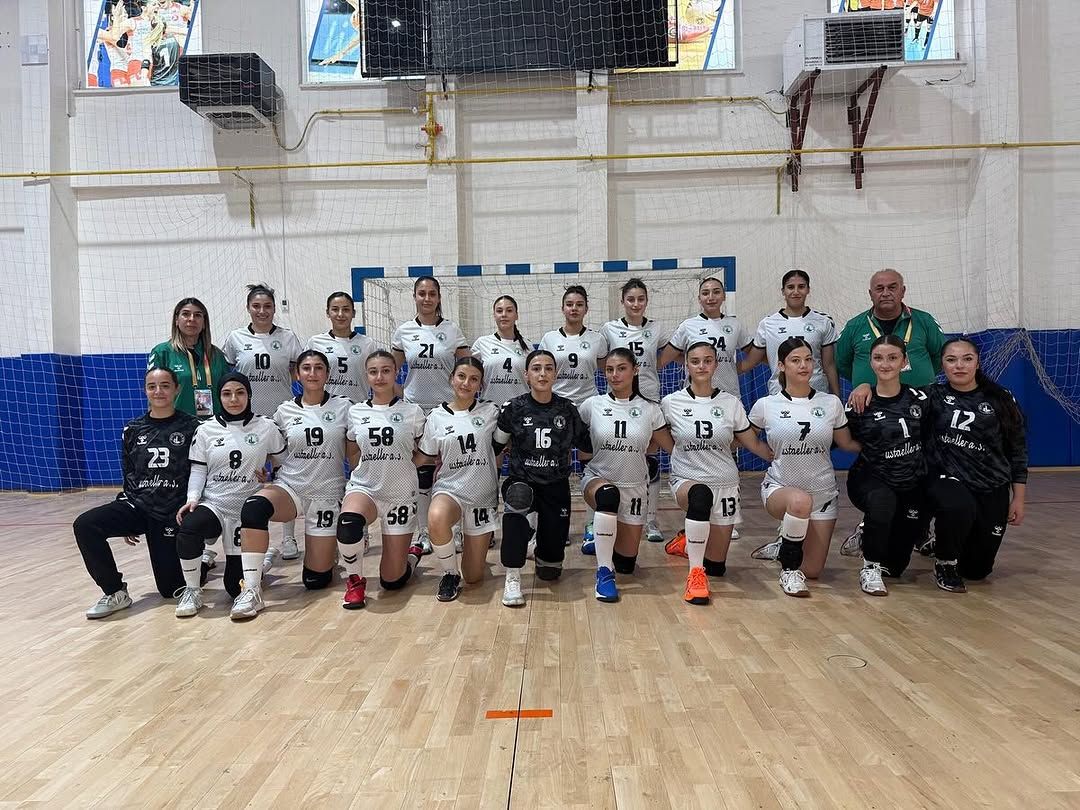 Spor salonunda, forma giyen kadın hentbolcular ve teknik ekip, kale önünde iki sıra halinde poz veriyor.