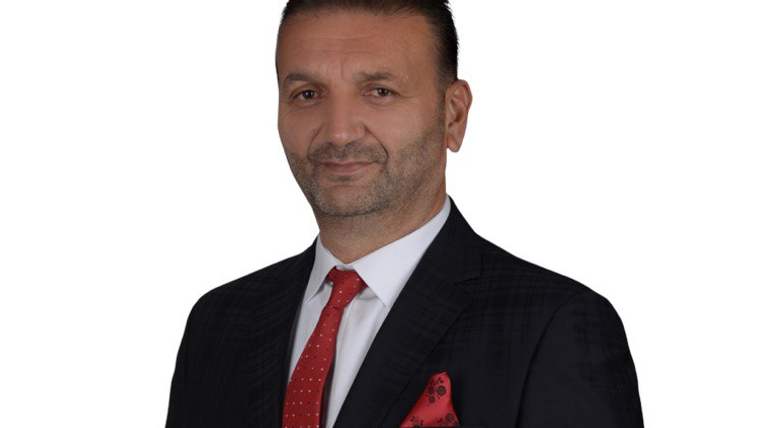 Beyaz arka plan önünde siyah takım elbise, kırmızı kravat ve mendil takan bir erkek portresi.