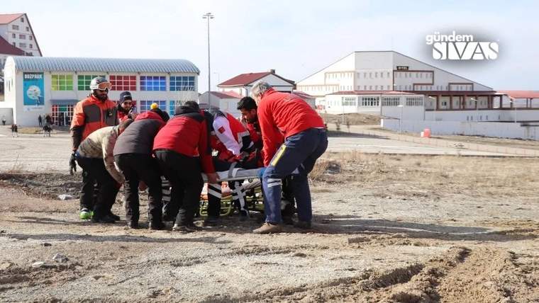 Açık alanda bir grup kişi, sedyede yatan kişiyi taşırken arka planda kayak merkezi binaları görülüyor.