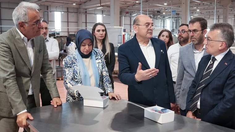Bir grup insan, bir fabrikanın içinde bir masanın etrafında toplanmış, konuşuyor.