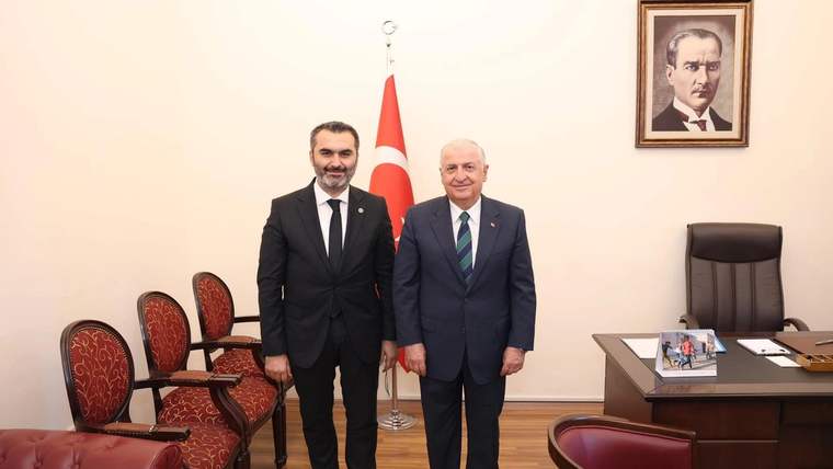 Türk bayrağı ve duvardaki portre önünde takım elbiseli iki kişi, bir makam odasında yan yana duruyor.
