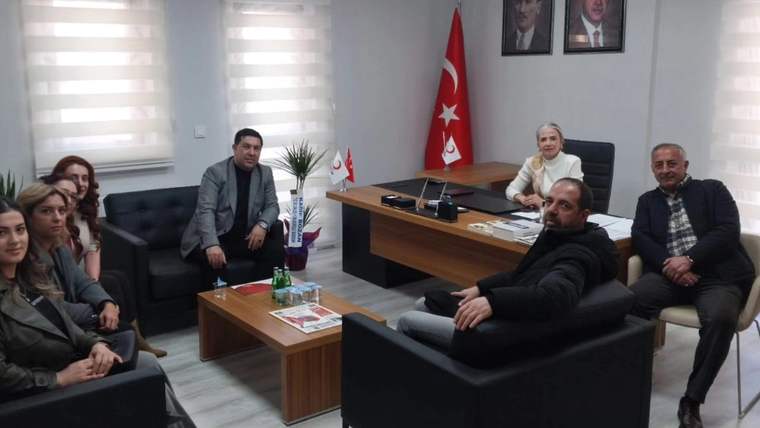 Ofis masası çevresinde oturan birkaç yetişkin, Türk bayrağı ve duvarlardaki portrelerin bulunduğu odada görülüyor.