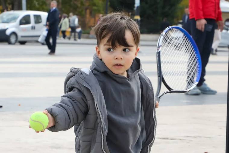 Açık alanda bir çocuk, elinde yeşil tenis topu ve raketle kameraya doğru duruyor.