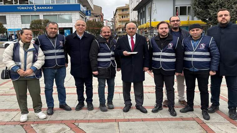 Kent meydanında, lacivert yelekli kişilerle birlikte duran bir kişi elindeki kâğıda bakıyor; arkada banka binası bulunuyor.
