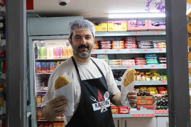 Sakallı bir kişi, kantin önünde iki ekmek arası yiyeceği ellerinde tutarken gülümsüyor.