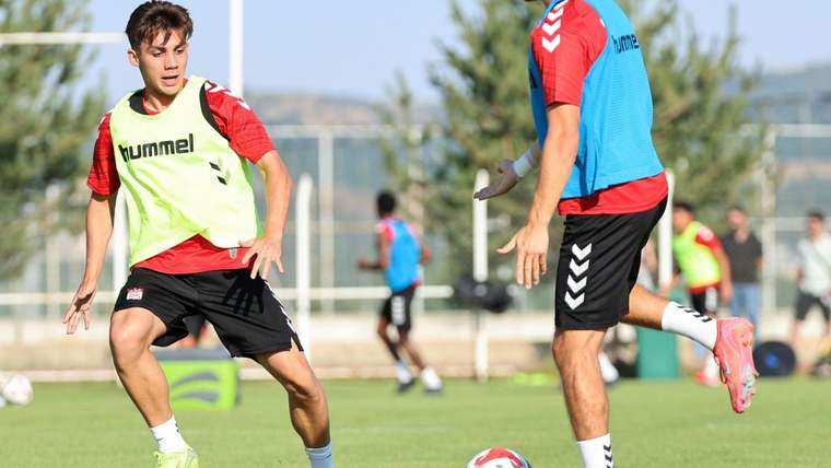 Çim sahada kırmızı-beyaz formalar giyen iki futbolcu, topun yanında antrenman yapıyor.