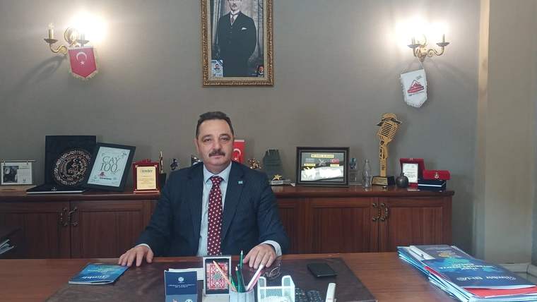 Bıyıklı bir kişi takım elbiseyle çalışma masasının arkasında oturuyor; duvarda portre ve çeşitli objeler bulunuyor.