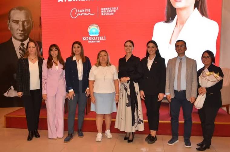 Kırmızı panolu salonda, dokuz kişi yan yana duruyor; arkalarında Atatürk portresi ve belediye logoları görülüyor.