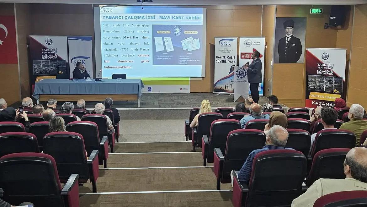 Bir salonda, SGK logosunun bulunduğu bir sunum ekranı önünde konuşan bir kişi ve dinleyiciler yer alıyor.