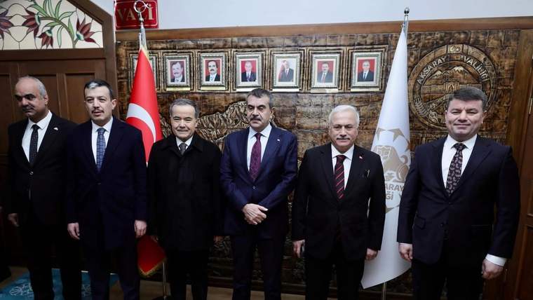 Ahşap panolar ve Türk bayrağı önünde, takım elbiseli beş erkek yan yana duruyor.