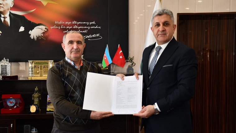 Ofis ortamında iki erkek, aralarında tuttukları imzalı belgeyle kameraya bakıyor; arkada Türk ve Azerbaycan bayrakları var.