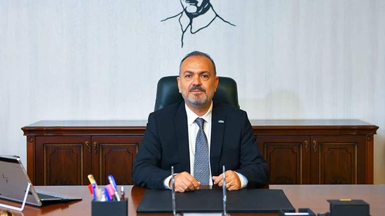 Takım elbiseli bir kişi, ahşap panelli bir odada masa başında oturuyor; önünde kalemlik ve mikrofonlar bulunuyor.