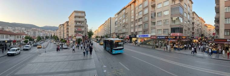 Geniş bir bulvarda turkuaz renkli belediye otobüsü, yayalar, araçlar ve çok katlı binalar görülüyor.