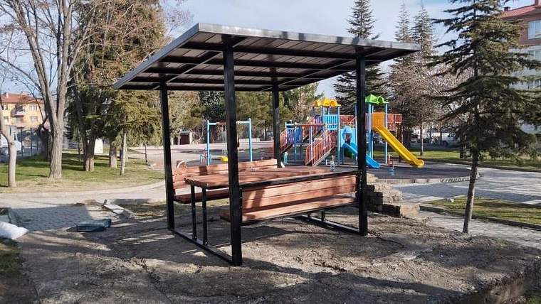 Önünde ahşap bank bulunan gölgelikli oturma alanı, arkasında kaydıraklı çocuk oyun grubuyla parkta yer alıyor.