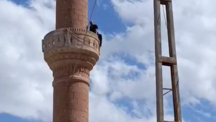 Bir kişi, taş minarenin şerefesinde dururken yanında metal bir merdiven yapısı görülüyor.