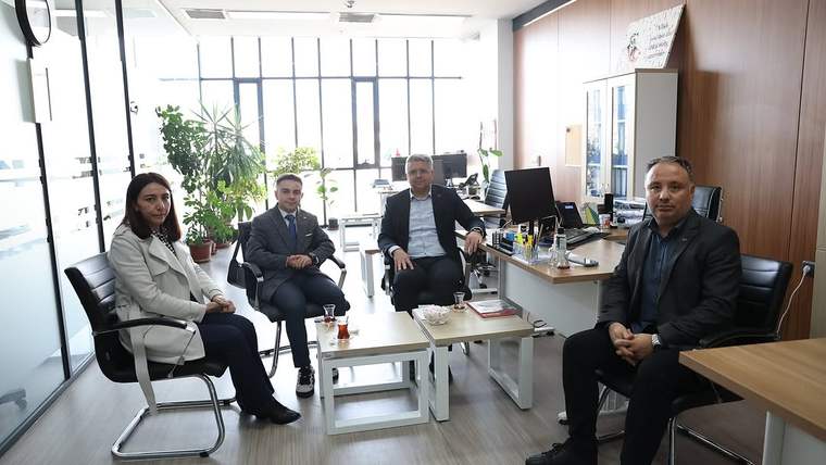 Ofis ortamında dört yetişkin, koltuk ve sandalyelerde otururken ortadaki sehpanın üzerinde çay bardakları bulunuyor.