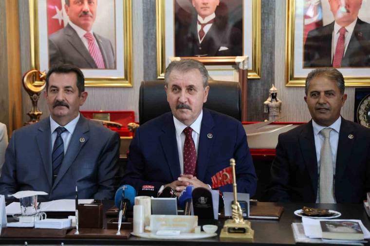 Mikrofonların bulunduğu masada ortada oturan bir kişi, iki kişinin arasında kameraya bakıyor.