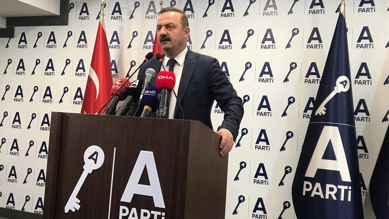 Kürsüde konuşan bir erkek, mikrofonların önünde duruyor; arkada A Parti logolu duvar ve bayraklar bulunuyor.