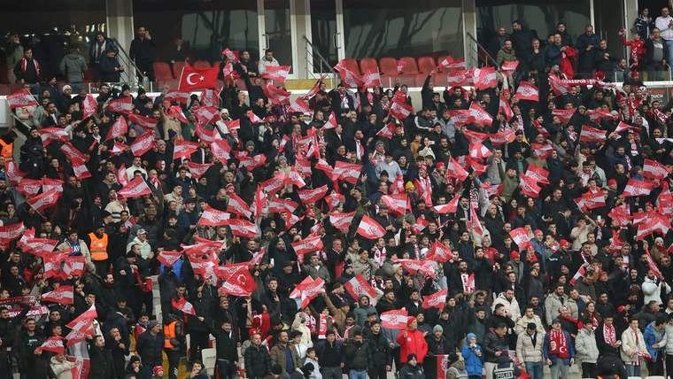 Tribünde çok sayıda taraftar, kırmızı-beyaz atkı ve bayraklarla birlikte ellerini kaldırıyor.
