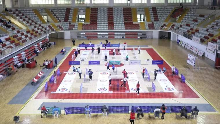 Bir spor salonunda floor curling turnuvası için hazırlık yapılıyor, oyuncular ve izleyiciler yer alıyor.
