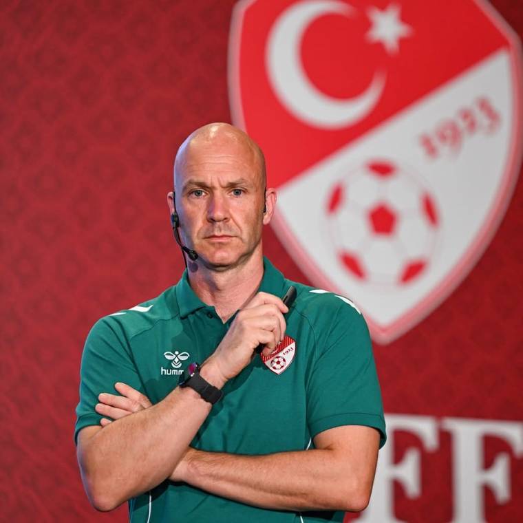 Kulaklık mikrofonu takan kel bir adam, yeşil polo tişörtüyle kollarını kavuşturmuş halde duruyor.