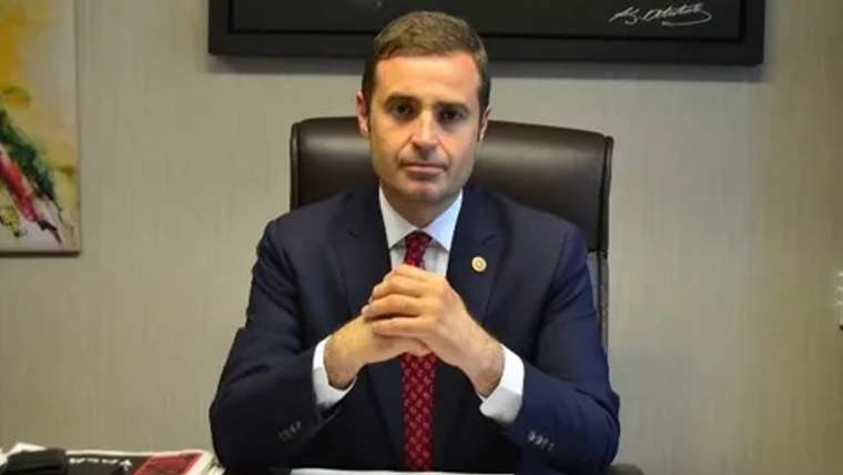 Bir masa başında oturan, takım elbise giymiş bir kişi, ellerini birleştirerek poz veriyor.