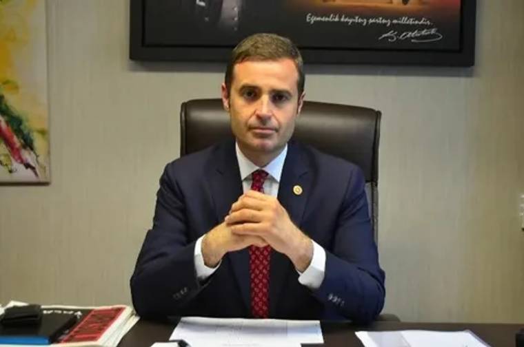 Bir masa başında oturan, takım elbise giymiş bir kişi, ellerini birleştirerek poz veriyor.
