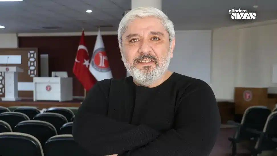 Kollarını kavuşturmuş, gülümseyen bir adam, arka planda Türk bayrağı ve bir kurum bayrağı görünmekte.