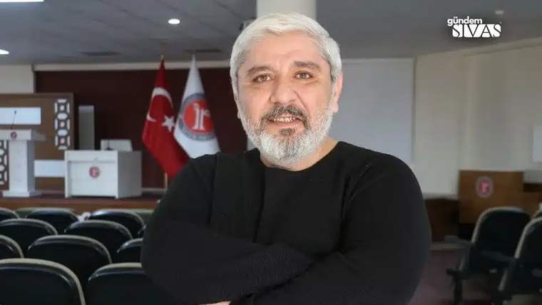 Kollarını kavuşturmuş, gülümseyen bir adam, arka planda Türk bayrağı ve bir kurum bayrağı görünmekte.