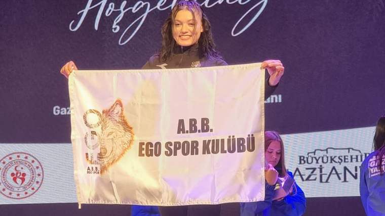 Bir kadın, A.B.B. EGO Spor Kulübü bayrağını tutarak sahnede gülümsüyor.