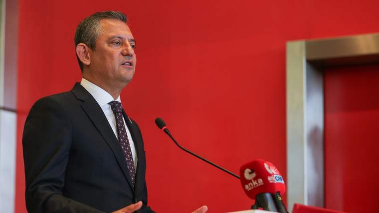Kırmızı duvar önünde takım elbiseli bir kişi, mikrofonların bulunduğu kürsüde konuşuyor.