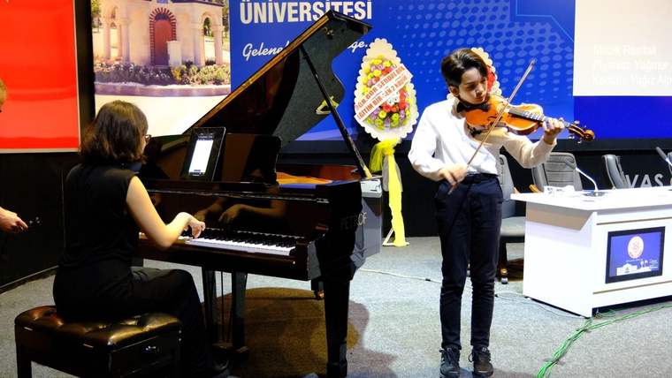 Piyano çalan bir kişi ile keman çalan bir kişi sahnede performans sergiliyor.