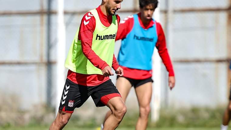 Yeşil ve mavi antrenman yeleği giyen iki futbolcu, açık sahada top sürerken görülüyor.