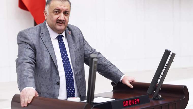 Takım elbiseli bir kişi, elektronik ekranlı ahşap kürsünün yanında durarak elini kürsüye dayıyor.