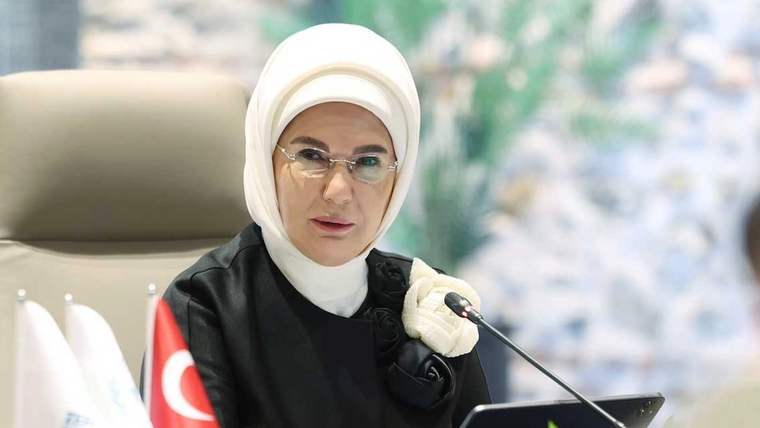 Kahverengi bir koltukta oturan bir kadın, beyaz başörtüsü ve gözlük takıyor.