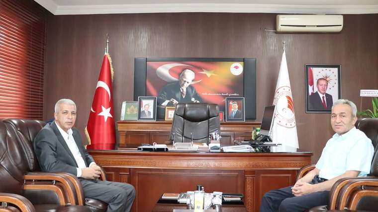 Ahşap panelli makam odasında, Türk bayrağı ve kurum flamaları önünde oturan iki kişi görülüyor.