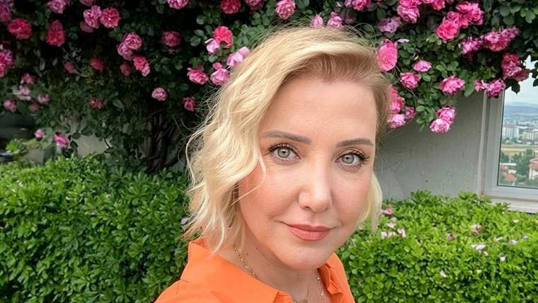 Turuncu gömlekli sarışın bir kadın, pembe çiçekli ve yeşil bitkilerle çevrili bahçede kameraya bakıyor.