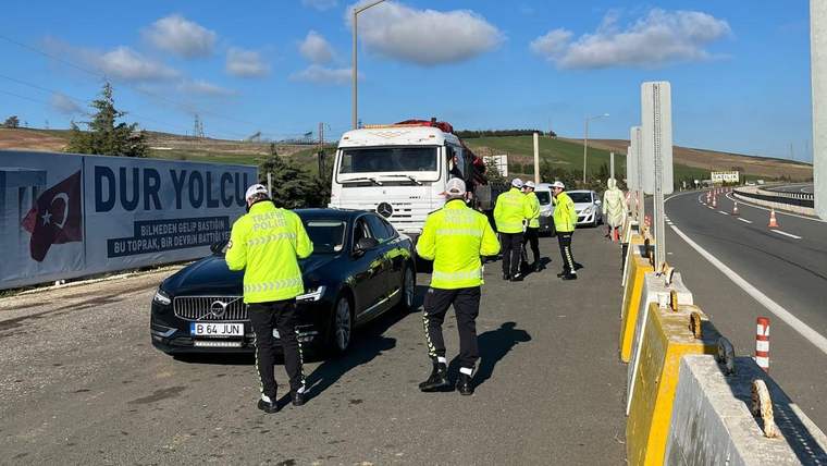 Reflektif yelekli trafik görevlileri, yol kenarında siyah otomobil ve beyaz kamyonu kontrol ediyor.