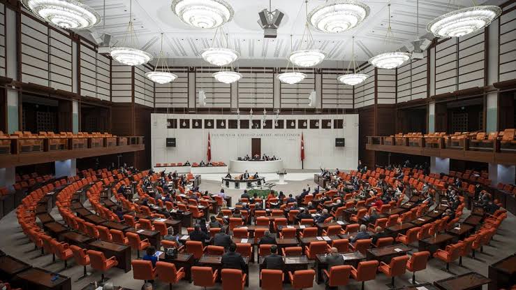Büyük bir meclis salonunda sıralarda oturan çok sayıda kişi ve önde kürsülerin bulunduğu genel görünüm.