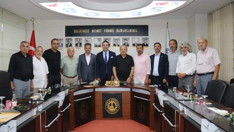 Toplantı salonunda, uzun masanın önünde yan yana duran bir grup insan ve iki Türk bayrağı görülüyor.