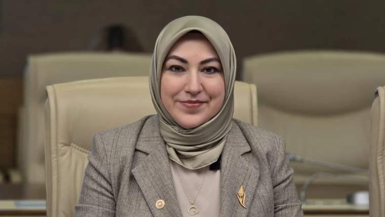 Başında başörtüsü olan bir kadın, gri ceket giymiş ve gülümseyerek kameraya bakıyor.