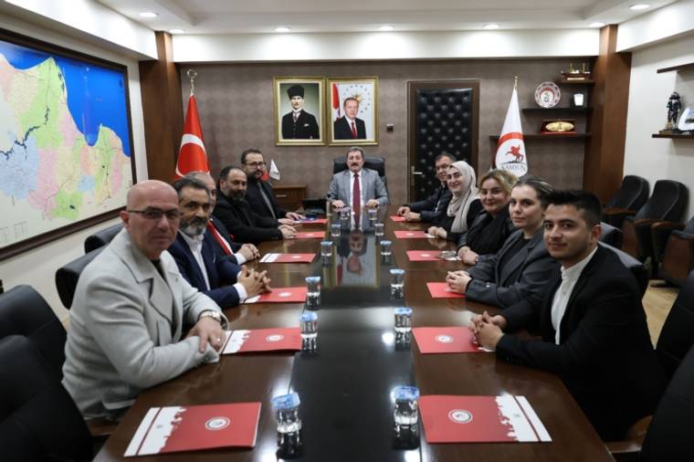 Toplantı odasında bir masa etrafında oturan on kişi, arka planda Türk bayrağı ve harita var.